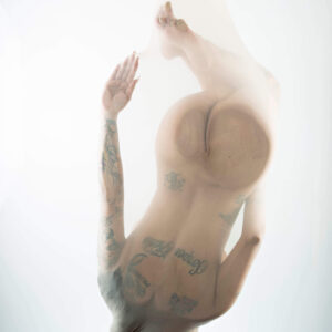 Maxi Soule poses curled upside down inside translucent white fabric, her tattooed body softly silhouetted against a bright white background.