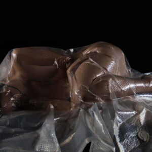Rebecca Lawrence lies curled on her side beneath a transparent plastic vacuum-seal sheet against a black background, with reflective folds contouring her body.