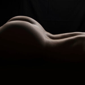 Rebecca Lawrence lies nude on her side against a black background, with low directional lighting tracing the curves of her back, hips, and legs.