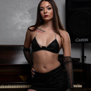 Shordy Kyy poses in a black two-piece outfit with sparkling sheer gloves in front of a vintage upright piano.