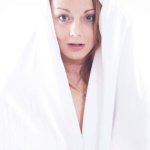 Close-up portrait of Bee Brunson looking directly at the camera while draped in flowing white fabric against a bright white studio background.