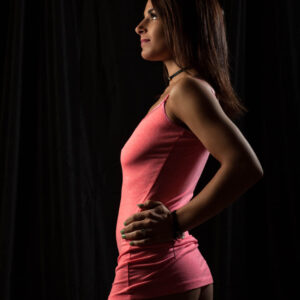 Tiffany K. poses in profile against a black backdrop wearing a fitted pink mini dress and black choker, with dramatic side lighting outlining her figure.