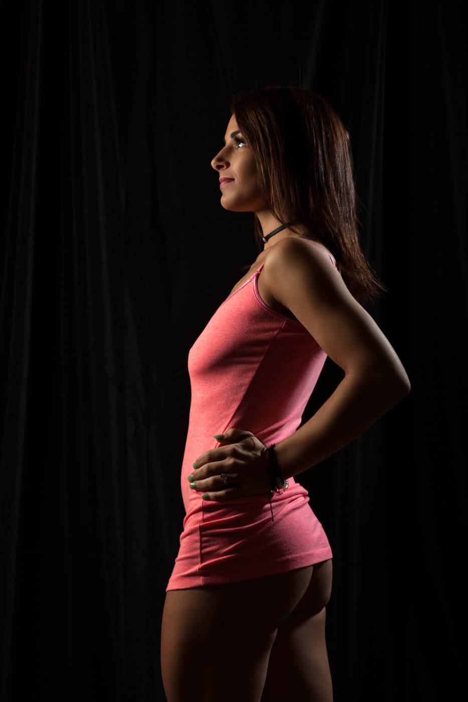Tiffany K. poses in profile against a black backdrop wearing a fitted pink mini dress and black choker, with dramatic side lighting outlining her figure.