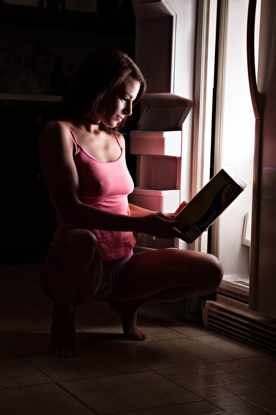 Tiffany K crouches beside an open refrigerator while reading a food box, illuminated primarily by the refrigerator light in a dark kitchen.