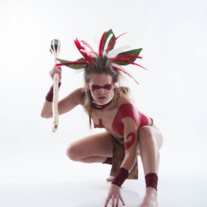 Shelby Wolf crouches in a dynamic pose against a bright white background, wearing red body paint, a dramatic red-and-white feathered headdress and holding a handmade staff.