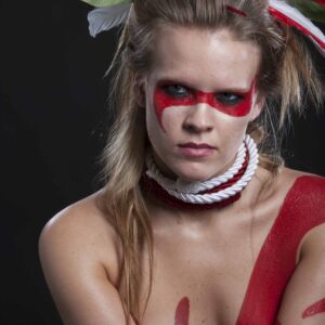 Shelby Wolf poses against a black background with dramatic red face and body paint, layered rope around her neck, and red, white and green feathers arranged through her hair.