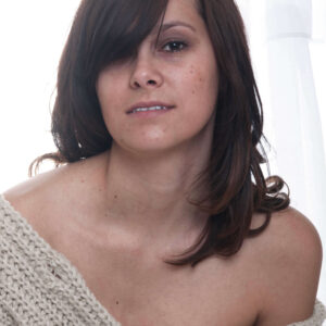 Close-up portrait of model Marisol L wearing a cream knit sweater draped off her shoulders, photographed against softly backlit white curtains.