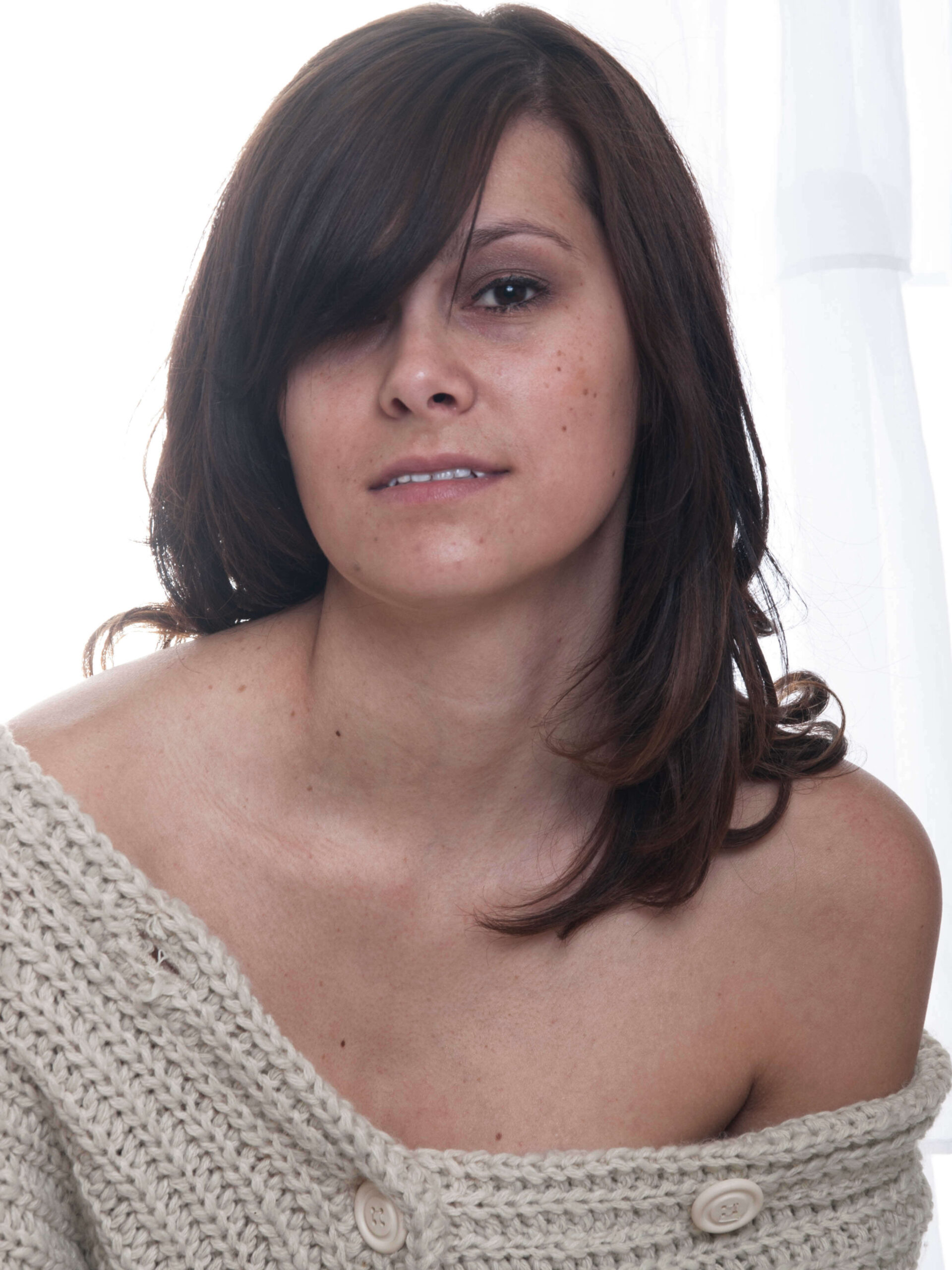 Close-up portrait of model Marisol L wearing a cream knit sweater draped off her shoulders, photographed against softly backlit white curtains.