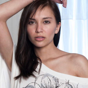 Model Kylie Hunter poses with one arm raised in a close-up fashion portrait, wearing an off-the-shoulder white floral graphic top.