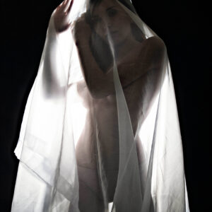 Model Kylie Hunter poses beneath translucent white fabric in a dark studio, with dramatic backlighting revealing her face and silhouette through the sheer material.