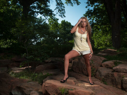 Traci Lacek poses with one foot on a rock in a shaded wooded landscape, wearing a pale yellow top, white shorts, and black high heels.