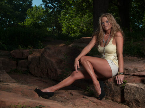 Traci Lacek sits on large rocks in a wooded setting with one leg extended, wearing a pale yellow top, white shorts, layered jewelry, and black heels.