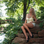 Traci Lacek sits on rocks beside a small pond in a lush garden, wearing a pale yellow sleeveless top, white shorts, jewelry, and black heels.