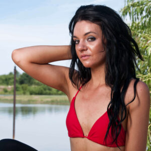 Rachel poses beside a pond wearing a red bikini top and black bottoms, resting one hand behind her head with willow branches framing the portrait.