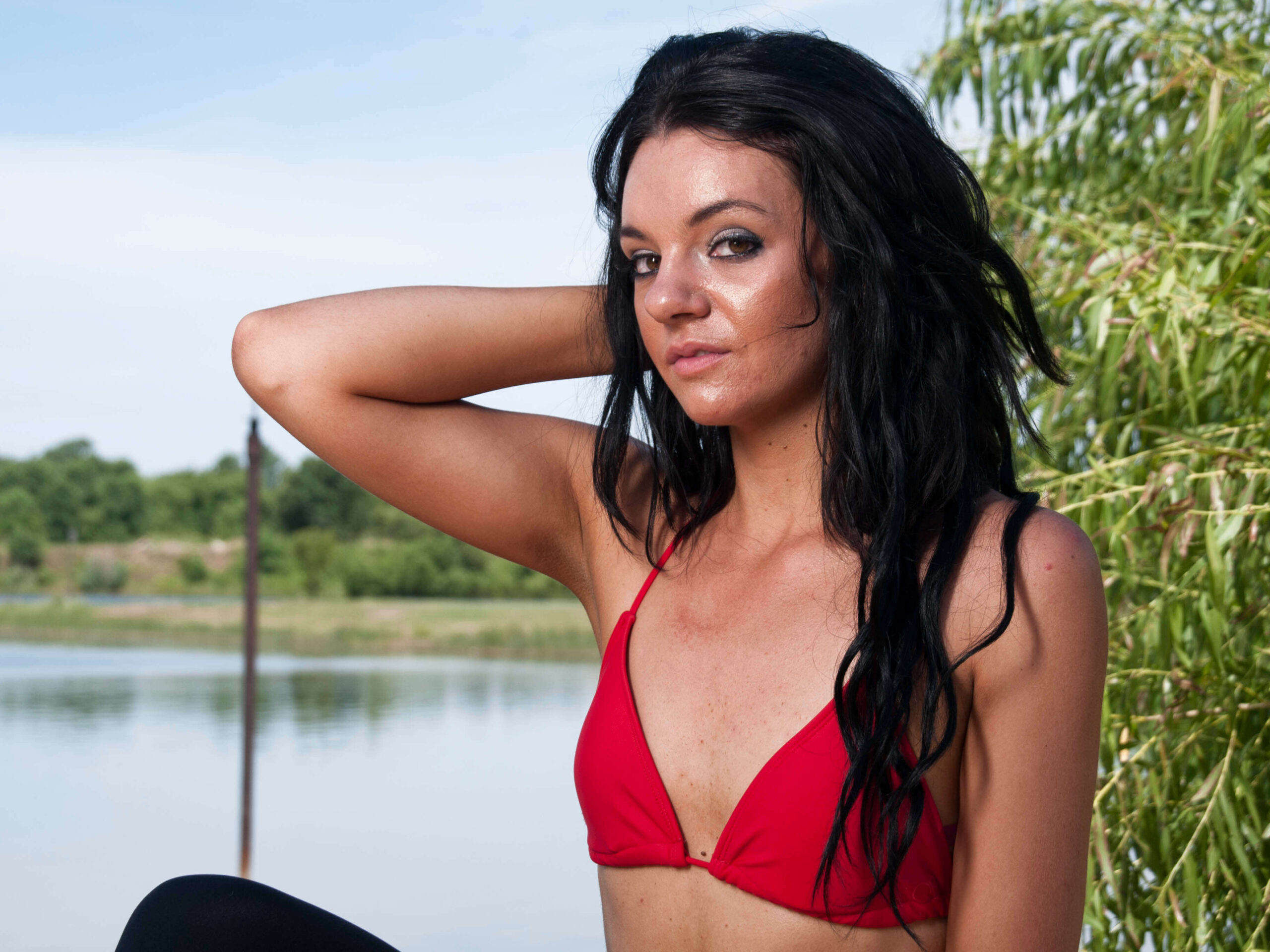 Rachel poses beside a pond wearing a red bikini top and black bottoms, resting one hand behind her head with willow branches framing the portrait.