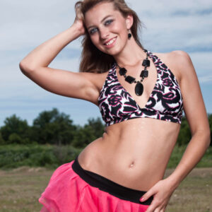 Outdoor glamour portrait of model Bree wearing a black, white, and pink patterned halter top, a bright pink layered skirt, and a bold black necklace while smiling in an open grassy field.