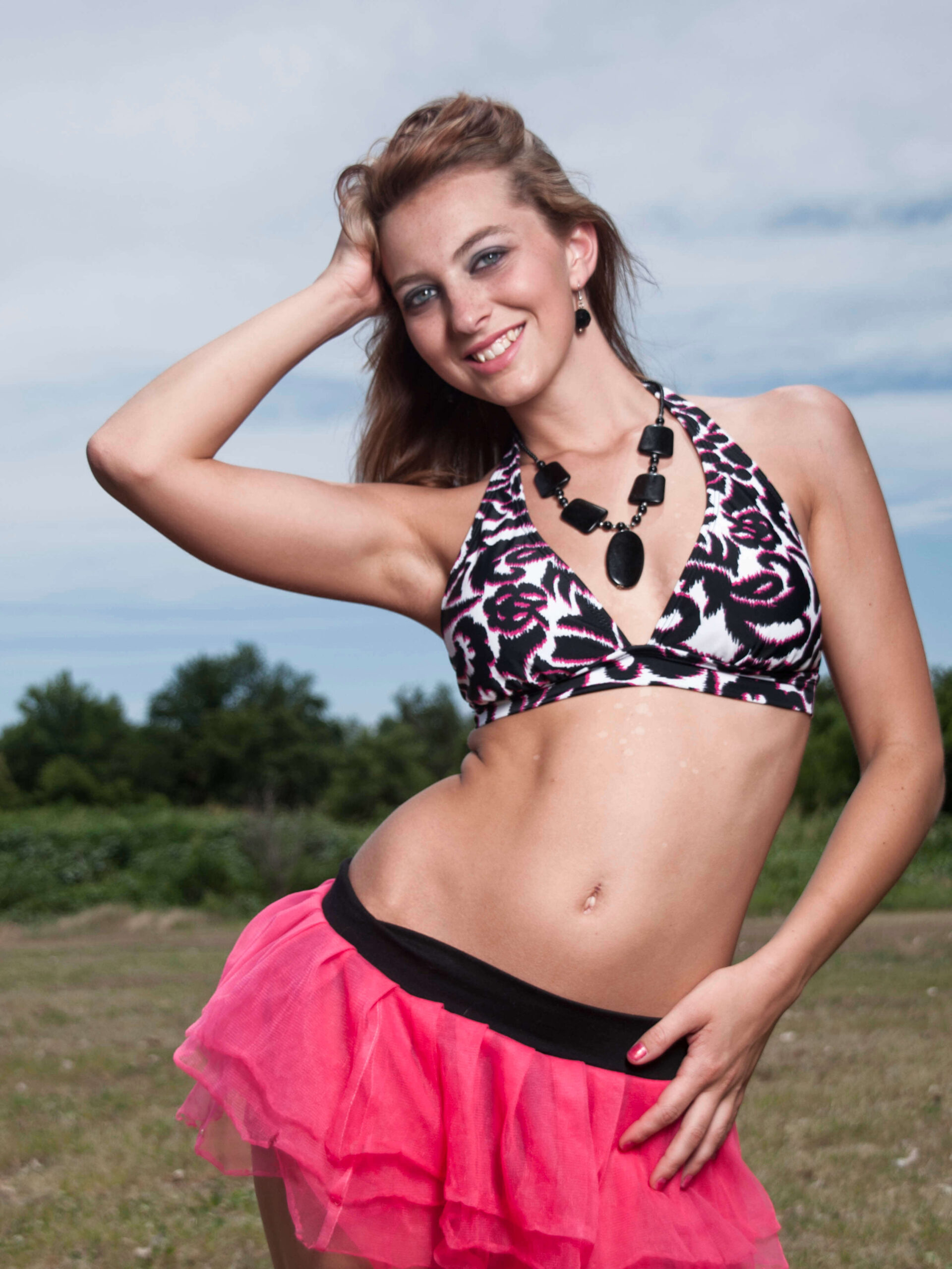 Outdoor glamour portrait of model Bree wearing a black, white, and pink patterned halter top, a bright pink layered skirt, and a bold black necklace while smiling in an open grassy field.