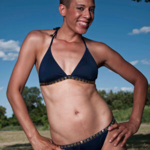 Jeanine Shay smiles while posing outdoors in a dark navy bikini beneath a vivid blue sky with trees and grass in the background.