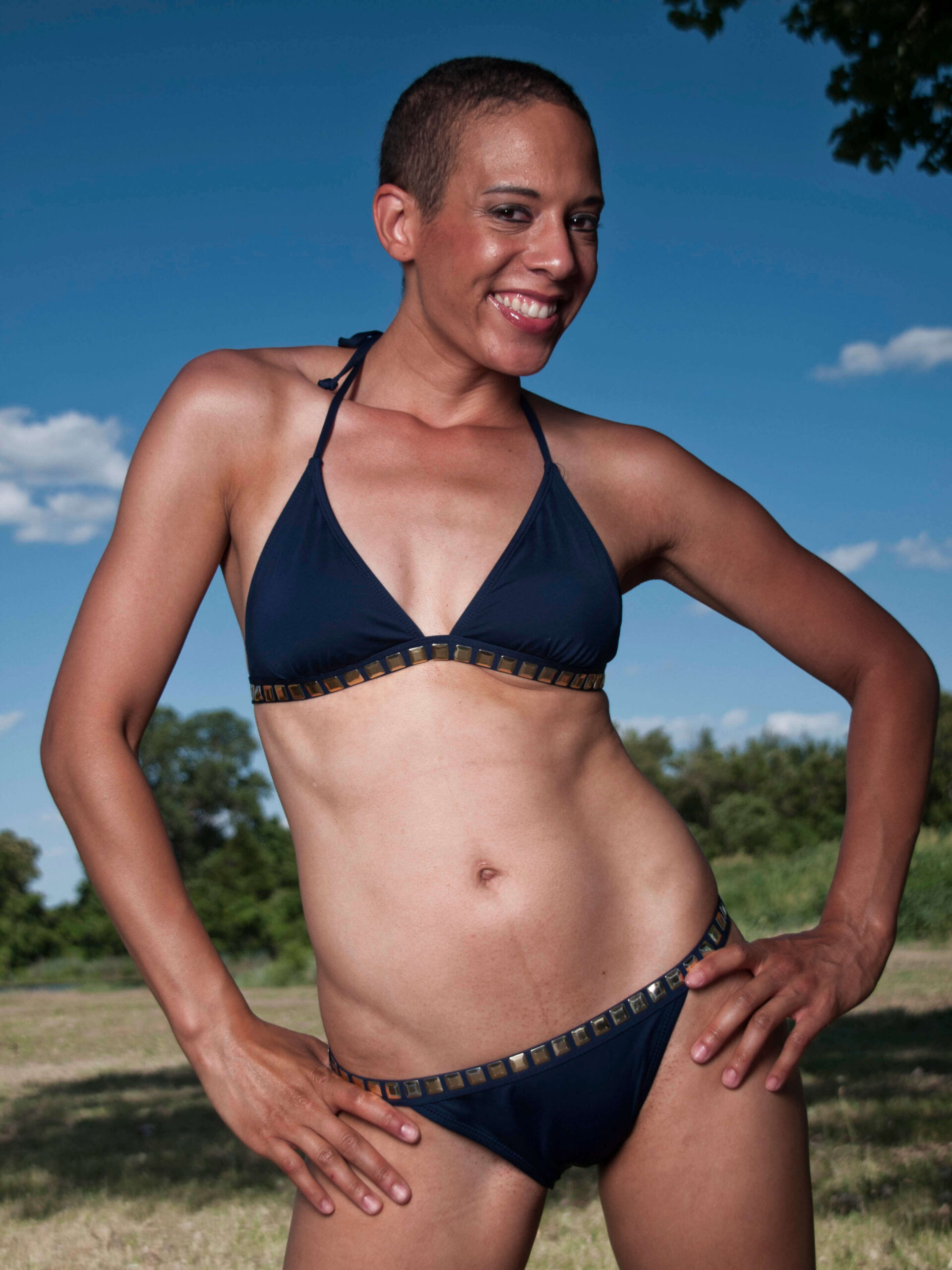 Jeanine Shay smiles while posing outdoors in a dark navy bikini beneath a vivid blue sky with trees and grass in the background.