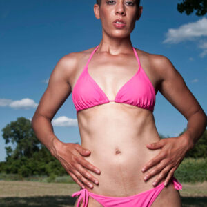 Jeanine Shay poses outdoors in a bright pink bikini, standing against a vivid blue sky with trees and an open grassy landscape behind her.