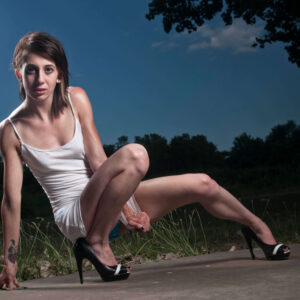 Tiffany poses outdoors at dusk in a white mini dress and black high heels, crouching dramatically on a concrete surface beneath a deep blue evening sky.