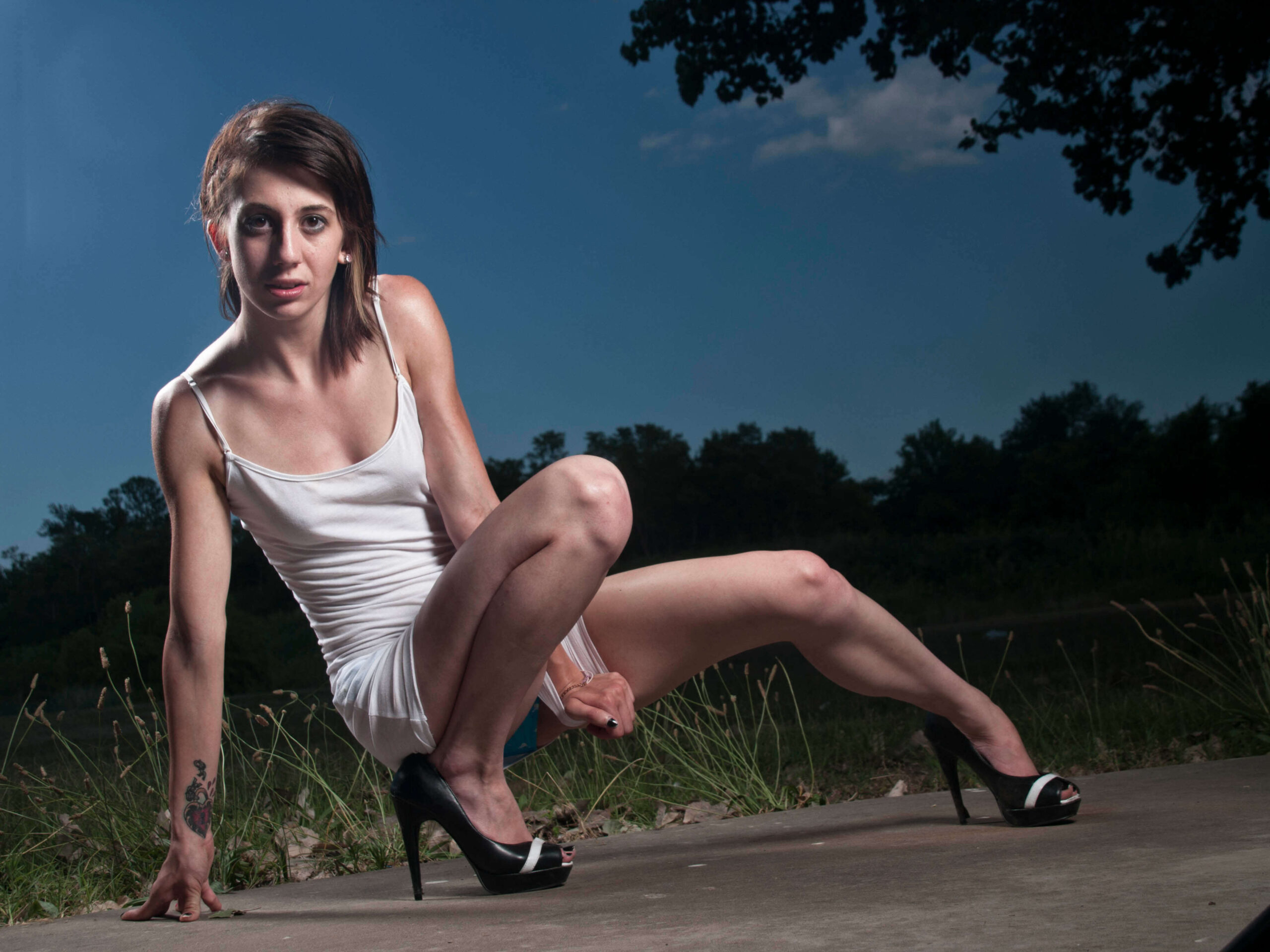 Tiffany poses outdoors at dusk in a white mini dress and black high heels, crouching dramatically on a concrete surface beneath a deep blue evening sky.