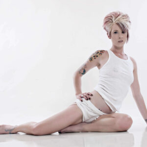 Tasha T. sits on a white studio floor wearing a white tank top and shorts, posing against a bright high-key background with her colorful arm tattoos visible.