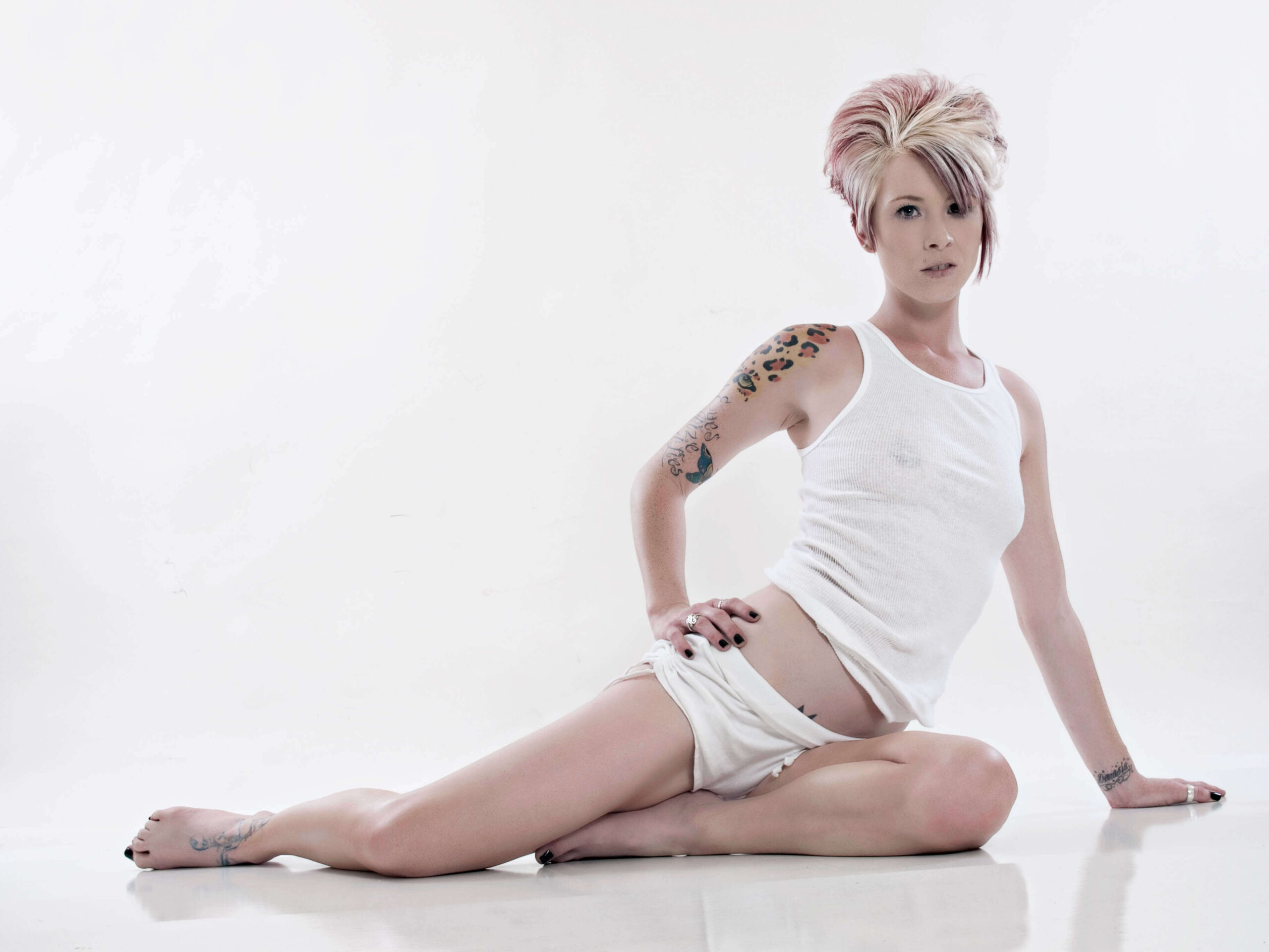 Tasha T. sits on a white studio floor wearing a white tank top and shorts, posing against a bright high-key background with her colorful arm tattoos visible.