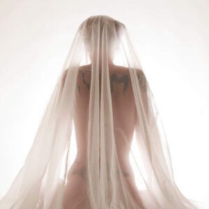 Tasha T. sits with her back to the camera beneath layers of sheer white fabric, her silhouette and tattoos softly visible through the translucent material against a bright background.