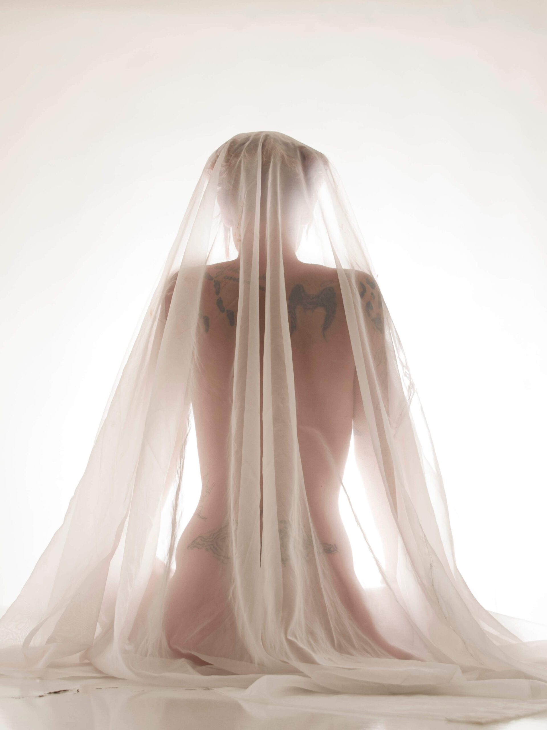 Tasha T. sits with her back to the camera beneath layers of sheer white fabric, her silhouette and tattoos softly visible through the translucent material against a bright background.