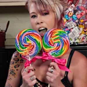 Tasha T poses playfully with two oversized rainbow swirl lollipops tied with bright pink bows, sticking out her tongue in a colorful candy-themed setting.