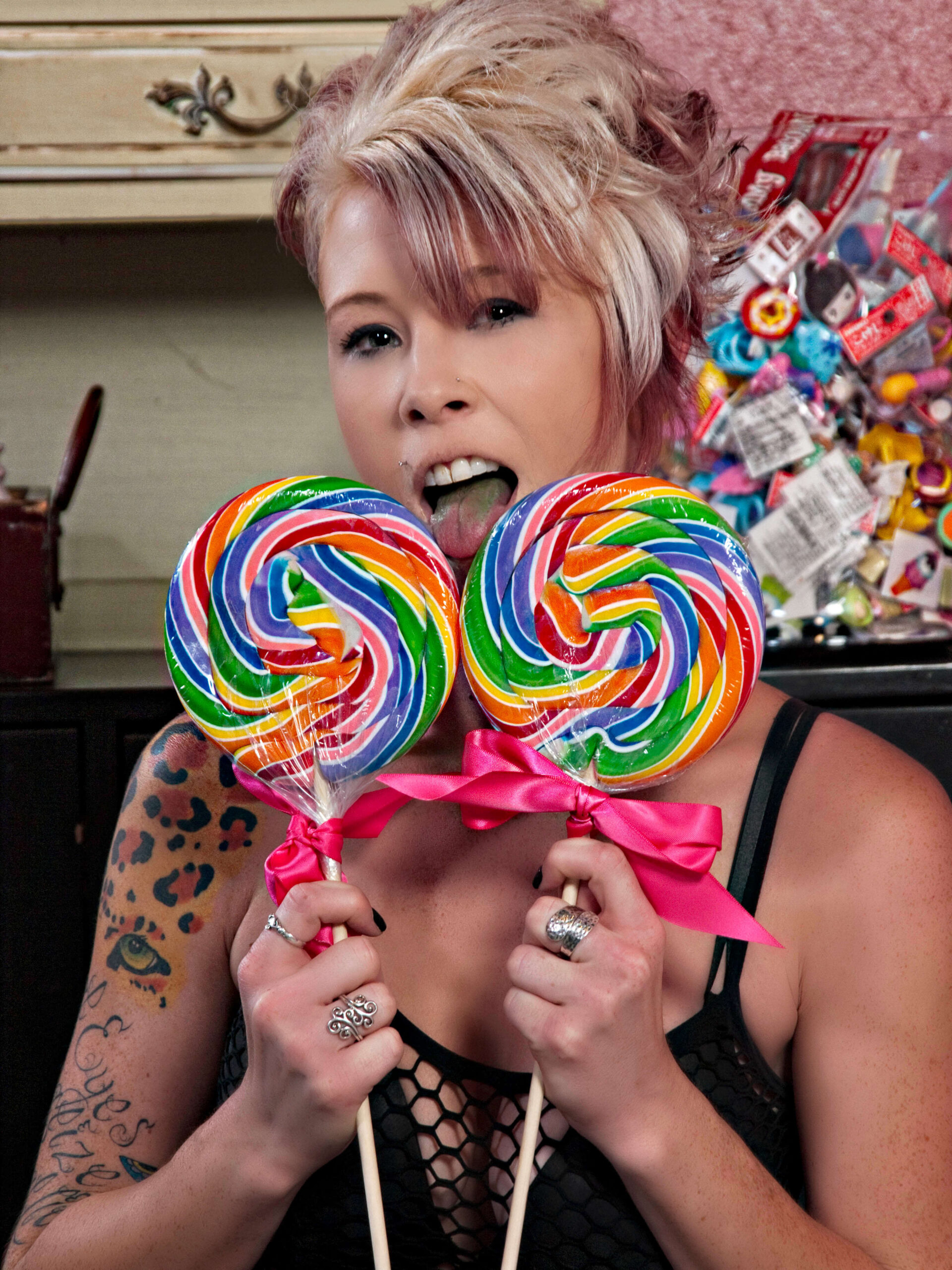 Tasha T poses playfully with two oversized rainbow swirl lollipops tied with bright pink bows, sticking out her tongue in a colorful candy-themed setting.