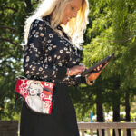 Sinderella stands outdoors beneath green trees, wearing a patterned black blouse and black skirt while using a tablet and carrying a red fashion handbag.