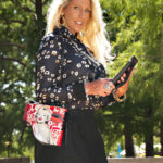 Sinderella looks toward the camera while holding a tablet outdoors, wearing a black patterned blouse, black skirt, statement jewelry, and a red handbag.