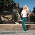 Sinderella poses in teal trousers and a sleeveless white top in front of a large cascading rock waterfall on a sunny day.