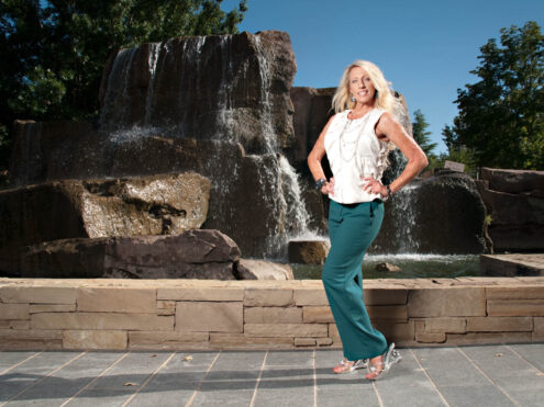 Sinderella poses in teal trousers and a sleeveless white top in front of a large cascading rock waterfall on a sunny day.