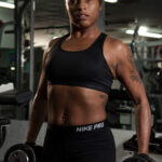 Athletic female fitness model wearing black Nike Pro workout clothing and holding dumbbells at her sides during a professional gym photoshoot.