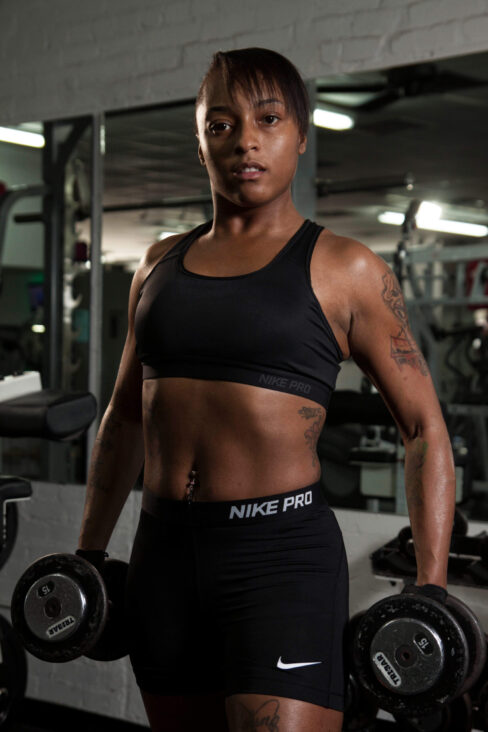 Athletic female fitness model wearing black Nike Pro workout clothing and holding dumbbells at her sides during a professional gym photoshoot.