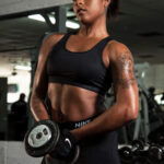 Female fitness model in black Nike Pro athletic wear holding dumbbells in front of her body during a strength training photoshoot inside a gym.