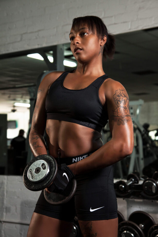 Female fitness model in black Nike Pro athletic wear holding dumbbells in front of her body during a strength training photoshoot inside a gym.