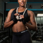 Smiling female fitness model wearing black Nike Pro athletic clothing and training gloves while holding a white workout rope around her neck inside a gym.