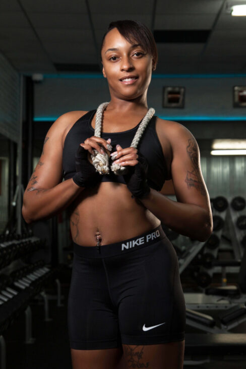 Smiling female fitness model wearing black Nike Pro athletic clothing and training gloves while holding a white workout rope around her neck inside a gym.