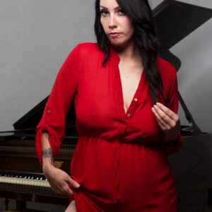 Felicity Sparks poses confidently in a vibrant red dress beside a vintage grand piano. Her dark flowing hair, alluring gaze, and subtly raised hemline create an elegant portrait blending sophistication with sensuality.