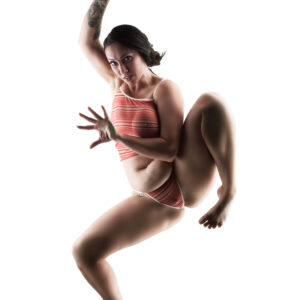 Felicity Sparks strikes a dynamic, gravity-defying pose against a brilliant white background. Wearing coral-and-white striped lingerie, she extends one tattooed arm overhead while raising a bent leg, creating an unconventional composition filled with movement and attitude.