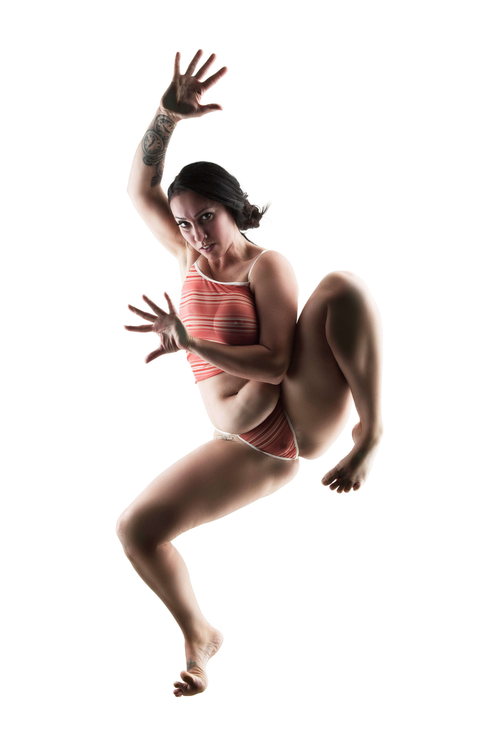 Felicity Sparks strikes a dynamic, gravity-defying pose against a brilliant white background. Wearing coral-and-white striped lingerie, she extends one tattooed arm overhead while raising a bent leg, creating an unconventional composition filled with movement and attitude.