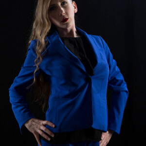 Hippy Researcher poses confidently in a vibrant royal blue pantsuit over a black blouse, her long blonde hair cascading over one shoulder against a dramatic black studio backdrop.
