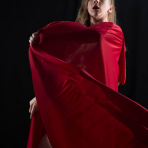 Hippy Researcher poses dramatically with flowing crimson-red fabric against a black studio backdrop, her long blonde hair and striking red lipstick complementing the vibrant material as it billows across the frame.