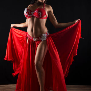 Madam Mayhem spreads a flowing red belly dance skirt while wearing a jeweled red costume against a black studio backdrop.