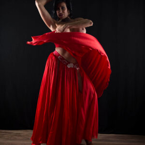 Madam Mayhem poses topless with one arm raised as flowing scarlet fabric sweeps across her body against a black studio backdrop.