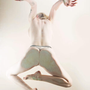 Hippy Researcher captured from behind in a dynamic airborne pose, wearing delicate light-colored underwear. Her arms extend upward and outward while her legs bend gracefully against a luminous white background, emphasizing movement and the sculptural beauty of the human form.